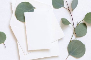 Kart ve zarf yeşil okaliptüs dalları yakınındaki beyaz bir masanın üzerinde ve yukarıdan görünüyor, Mockup. Minimum romantik daire ve boş yatay kart. Düğün ve parti kırtasiyesi
