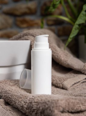 Kahverengi havluların üzerinde beyaz krem pompa şişesi ve banyoda yeşil bitki, yakın çekim, kozmetik ambalaj modeli. Kişisel bakım için doğal yüz kremi