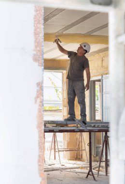 Caftsman bir odanın içinde iskelede tavana kadar çalışıyor. İnşaat veya yenileme çalışmaları sırasında tozlu gri iş kıyafetleri ve beyaz kasklı bir adam.