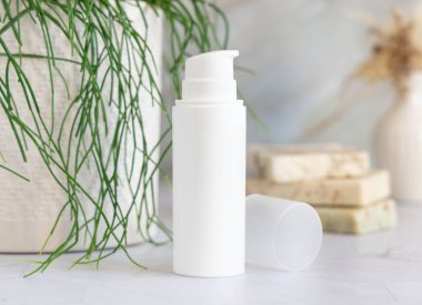 Banyodaki yeşil saksı bitkisinin yanındaki beyaz mermer tezgahta beyaz krem pompa şişesi. Kapatın, ambalaj modeli. Yüz veya vücut bakımı için doğal kozmetik ürünü