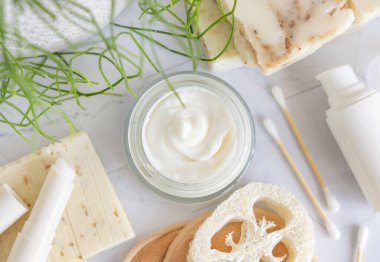 Yeşil bitkinin yanında beyaz mermer üzerine açık krema kavanozu ve banyo manzaralı sabun barları, ambalaj modeli. Yüz bakımı için doğal kozmetik ürünler, güzellik rutini