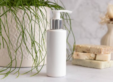 Banyodaki yeşil saksı bitkisinin yanındaki beyaz mermer tezgahta beyaz krem pompa şişesi. Kapatın, ambalaj modeli. Yüz veya vücut bakımı için doğal kozmetik ürünü