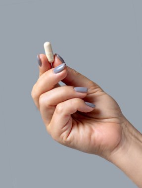 Gri arka planda parmakları yakın plan olan bej bir kapsülü tutan bir kadın el. Sağlık, Tıbbi ve İlaç Konsepti