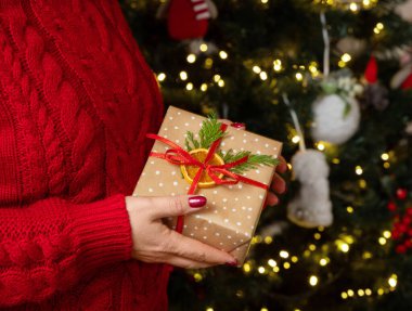 Kırmızı kazaklı bir kadın Noel ağacı ışıklarına karşı ellerini uzatmış, yandan bakıyor. Kırmızı tırnaklı kız hediye paketi saklıyor. Sıcak tatil ortamı