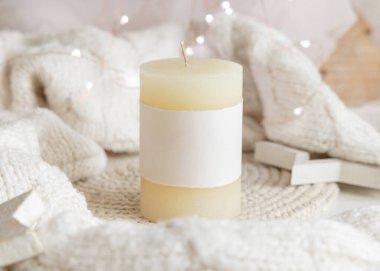 Beyaz örgü süveterin yanında boş etiketli sütun mumu, ahşap Noel dekoru ve ışıklar, kopyalama alanı. Kış atmosferi kırsal ya da İskandinav stiliyle benzer.