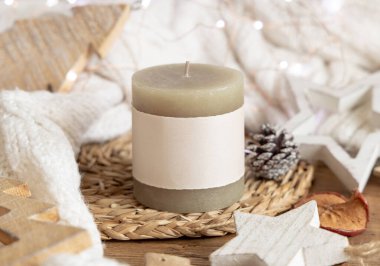 Beyaz örgü süveterin yanında boş etiketli sütun mumu, ahşap Noel dekoru ve ışıklar, kopyalama alanı. Kış atmosferi kırsal ya da İskandinav stiliyle benzer.