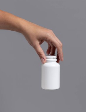 Beyaz plastik bir hap şişesi tutan kadın el gri arka plana yakın plan. Hapların ya da bitkisel ilacın için ambalaj modeli.