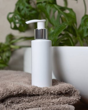 Kahverengi havlunun üzerinde beyaz krem pompa şişesi ve banyoda yeşil bitki, yakın çekim, kozmetik ambalaj modeli. Kişisel bakım için doğal erkek yüz kremi