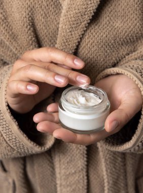 Kahverengi bornozlu, kreme dokunan ve elinde açık kozmetik kavanozu tutan kadının yakın plan fotoğrafı, doğal manikür. Vücut ve yüz bakımı için organik kozmetik ürünleri