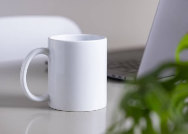 Gri ofis masasındaki 11 ons beyaz kahve fincanını açık dizüstü bilgisayar, gözlük ve yeşil bitki maketine yakın çekim. Kopyalama alanı olan tasarım şablonu