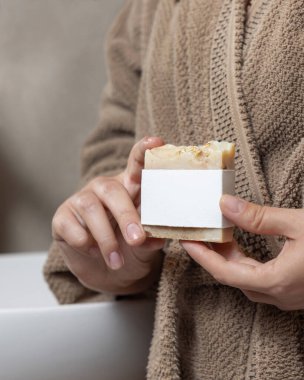 Female hands with natural manicure holding soap bar with blank label, packaging mockup. Close up of woman in bathroom wearing light brown bathrobe. Hygiene concept clipart