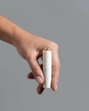 Beyaz dudak kremi çubuğunu gri renge yakın, kozmetik paket maketine yapıştıran kadın el, kopyalama alanı. Minimum bileşimli dudak bakımı güzellik ürünü