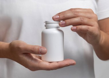 Beyaz tişörtlü, elinde beyaz plastik ilaç şişesi olan bir kadın. Bitkisel takviye için ambalaj modeli. Doğal manikürlü kadın elleri.