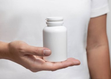 Beyaz tişörtlü, elinde beyaz plastik ilaç şişesi olan bir kadın. Bitkisel takviye için ambalaj modeli. Doğal manikürlü kadın elleri.