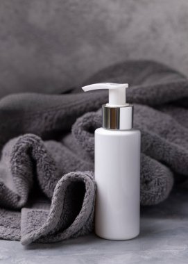 Banyoda koyu gri havlunun yanında beyaz boş bir kozmetik şişesi, yakın plan, marka ambalaj modeli, kopyalama alanı. Yüz bakımı ve spa prosedürleri, losyon veya toner için doğal el yapımı ürünler