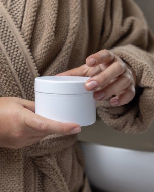 Doğal manikürlü, beyaz boş krema kavanozu tutan kadın elleri, kozmetik modelleme. Banyoda açık kahverengi bornoz giyen bir kadın. Bakım konsepti