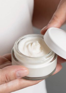 Beyaz gömlek ve gri arka planda krema kavanozu açan kadın, parmakları doğal manikürlü. Vücut veya yüz bakımı için organik kozmetik kavramı, günlük güzellik rutini