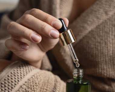 Doğal manikürlü kadın elleri beyaz yeşil kozmetik şişesini açıyor. Banyodaki kadın açık kahverengi bornoz giyiyor ve pipetten bir damla serum veya özü dökülüyor.