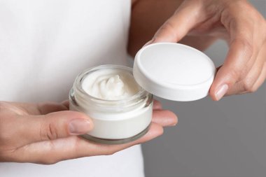 Beyaz tişört ve gri arka planda krem kavanozu açan kadın, doğal manikürlü parmaklar, kozmetik modelleme. Vücut veya yüz bakımı için organik kozmetik kavramı