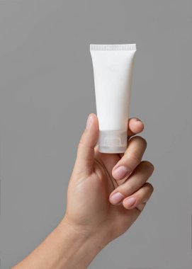 Beyaz sıkma krem tüpünü, gri kaplama kozmetik paket modellemesine karşı tutan kadın el, kopyalama alanı. En az bileşime sahip cilt bakımı ürünü