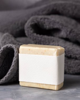 Koyu gri katlı havlunun yanında boş etiketli bej el yapımı sabun çubuğu, ambalaj modeli, kopyalama alanı. Günlük temizlik ve cilt bakımı için doğal bitkisel sabun.