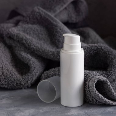 Banyodaki koyu gri havlunun yanında beyaz boş bir pompa krem şişesi, marka ambalaj modeli, kopyalama alanı. Yüz bakımı ve spa işlemleri için doğal el yapımı kozmetik ürünü