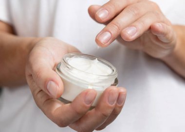 Close up of woman touching cream with finger and holding a cosmetic jar in other hand with natural nanicure agains white shirt. Concept of organic cosmetics for body care clipart