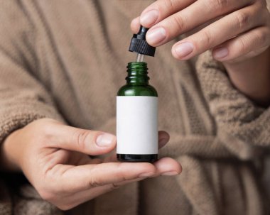 Doğal manikürlü kadın elleri yeşil kozmetik şişesini açıyor, etiket modeli. Açık kahverengi bornoz giyen bir kadının serum ya da özü kullandığına yakın çekim.