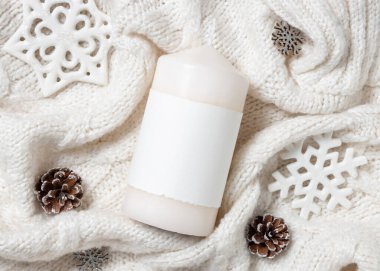 Kar taneleri ve çam kozalaklarının yanında beyaz örgü süveter üzerine boş etiketli beyaz sütun mumu, kopyalama alanı. Sıcak kış atmosferik modelleme, büyülü tatil zamanı. Noel ya da Yeni Yıl Selamlaması