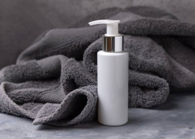 Banyoda koyu gri havlunun yanında beyaz boş bir kozmetik şişesi, yakın plan, marka ambalaj modeli, kopyalama alanı. Yüz bakımı ve spa prosedürleri, losyon veya toner için doğal el yapımı ürünler