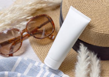 Havlunun yanındaki rattan şapkalı beyaz boş krema tüpü, kurutulmuş çimenler ve yakın plan güneş gözlüğü. Güneş kremi kozmetikleri