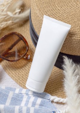Havlunun yanındaki rattan şapkalı beyaz boş krema tüpü, kurutulmuş çimenler ve yakın plan güneş gözlüğü. Güneş kremi kozmetikleri