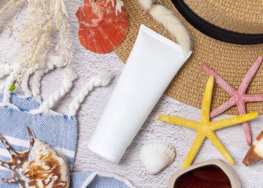 Havlunun, deniz kabuklarının ve deniz yıldızlarının yanındaki şapka torbasının üzerinde beyaz, boş bir krem tüpü var. Güneş kremi kozmetikleri