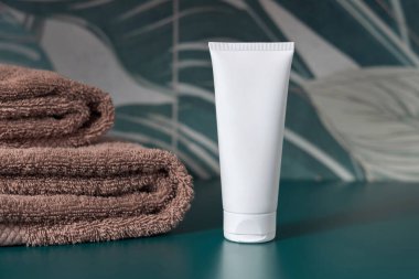 Kahverengi katlanmış havluların yanındaki beyaz krem sıkma tüpü koyu yeşil tropikal banyoya yakın, ambalaj modeli. Doğal bitki markasız kozmetik kabı, kopyalama uzayı. Spa ve sağlık konsepti