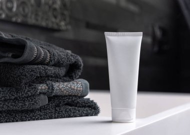 Siyah katlanmış havluların yanında beyaz krem sıkma tüpü koyu gri erkek banyosunda, yakın çekim, ambalaj modeli. Fotokopi alanı ile markasız doğal bitki kozmetikleri. Spa ve sağlık konsepti