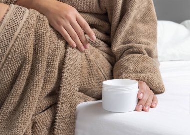 Kahverengi bornozlu kadın beyaz açık krema kavanozunu beyaz çarşafın üzerine koyuyor. Bakım konsepti. Vücut ve yüz bakımı için organik kozmetik ürünleri, güzellik ve kaplıca prosedürleri