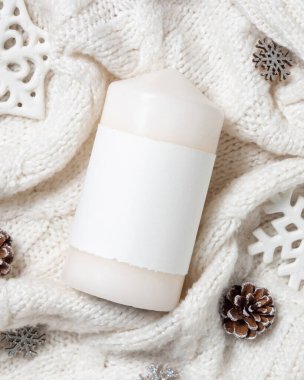 Kar taneleri ve çam kozalaklarının yanında beyaz örgü süveter üzerine boş etiketli beyaz sütun mumu, kopyalama alanı. Sıcak kış atmosferik modelleme, büyülü tatil zamanı. Noel ya da Yeni Yıl Selamlaması