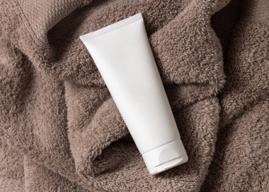 Banyonun üst görüntüsünde kahverengi havlunun üzerinde beyaz, boş krem tüpü, marka ambalaj modeli, kopyalama alanı. Yüz bakımı ve spa işlemleri için doğal el yapımı kozmetik ürünü