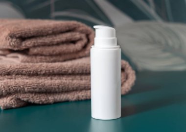 Kahverengi katlanmış havluların yanındaki beyaz krema şişesi koyu yeşil tropikal banyoda kapanıyor, ambalaj modeli. Doğal bitki markasız kozmetik kabı, kopyalama uzayı. Spa ve sağlık konsepti