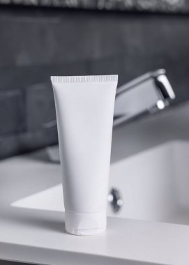 Modern musluğun yanındaki beyaz krem tüpü koyu gri, erkeksi tuvalette kapanıyor, ambalaj modeli. Kopya alanı olan doğal markasız kozmetik ürünler. Spa ve sağlık konsepti