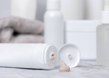 Açık kapaklı beyaz krema tüpü ve beyaz mermer masaya sıkıştırılmış bej krem, kozmetik şişeler ve gri banyo havlusuna karşı, yaklaşın, maket yapın. Günlük yüz bakımı için doğal ürün, spa prosedürleri