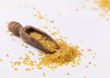 Çiğ, çiğ, sarı bulgur tanecikleri beyaz masadaki ahşap bir kepçe içinde yakın plan. Orta Doğu kuzeni. Sağlıklı beslenme için organik süper yiyecekler. Eski alternatif mısır gevreği. tohumlar