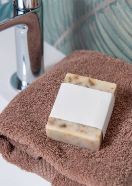 Yeşil tropikal banyodaki leğenin üzerindeki kahverengi katlanmış havluların üzerinde boş etiketli bej sabun kalıbı, fotokopi alanı ile paketleme. Günlük hijyen için markasız doğal bitki ürünü.