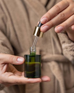 Doğal manikürlü kadın elleri beyaz yeşil kozmetik şişesini açıyor. Banyodaki kadın açık kahverengi bornoz giyiyor ve pipetten bir damla serum veya özü dökülüyor.