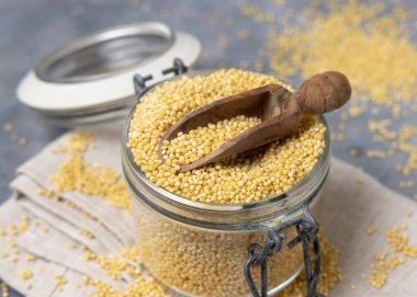 Cam kavanoz çiğ sarı darı taneciği. Gri masa üzerinde ahşap bir kepçe var. Yakın plan, kopyalama alanı. Eski psödocereal tohumlar. Organik süper yiyecekler. Sağlıklı gıda konsepti. Glütensiz alternatif tohumlar