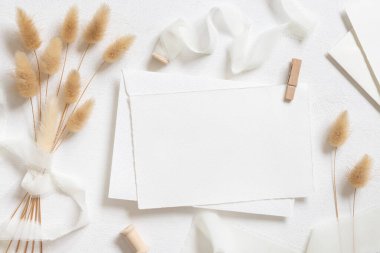 Beyaz kart ve zarf kıvrımlı ipek kurdelelerin yanında ve kurutulmuş tavşanın kuyruk çim üst görünümü beyaz, maket. Pastel romantik düz yatak. Parti tasarımı ya da düğün kırtasiyesi için bohem ya da doğa kartlarına yakın