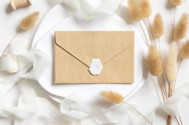 Kıvrımlı ipek kurdelelerin yanında mühürlü boş bir zarf ve kurutulmuş tavşanın kuyruk çim üst görünümü beyaz, maket. Pastel romantik düz yatak. Parti tasarımı ya da düğün kırtasiyesi için bohem ya da doğa kartlarına yakın