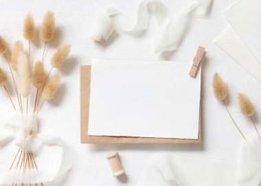 Beyaz kart ve zarf kıvrımlı ipek kurdelelerin yanında ve kurutulmuş tavşanın kuyruk çim üst görünümü beyaz, maket. Pastel romantik düz yatak. Parti tasarımı ya da düğün kırtasiyesi için bohem ya da doğa kartlarına yakın