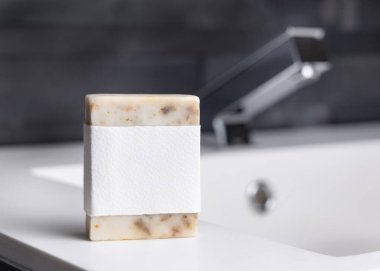 Modern koyu gri erkek banyosunda lavabonun yanında boş etiketli bej el yapımı sabun kalıbı, maket. Günlük erkek hijyeni ve vücut bakımı için doğal bitkisel ürünler