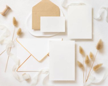 Bukleli ipek kurdelelerin yanında boş kartlar ve zarflar ve kurutulmuş tavşanın kuyruk çimlerini beyazın üstünde, maket. Pastel romantik düz yatak. Parti tasarımı ya da düğün kırtasiyesi için bohem ya da doğa kartlarına yakın